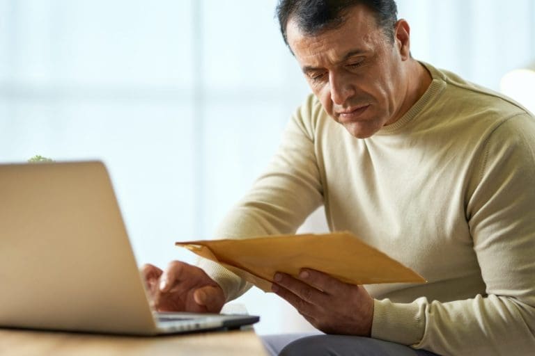 Portrait of busy latin middle aged businessman looking at the envelope, using laptop while working